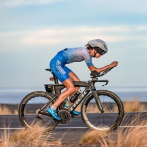 Rachel Zilinskas rides her black Trek TT bike on the Queen K in Kona, Hawaii in 2023. She is wearing a blue and white kit with a white aero helmet and is in her aero bars.