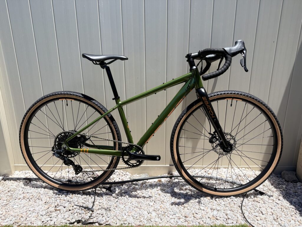 Batch GB.1 gravel bike shown from the non-drive side, olive-green aluminum frame with copper accents and tan-wall gravel tires, leaning against a light-panelled wall.